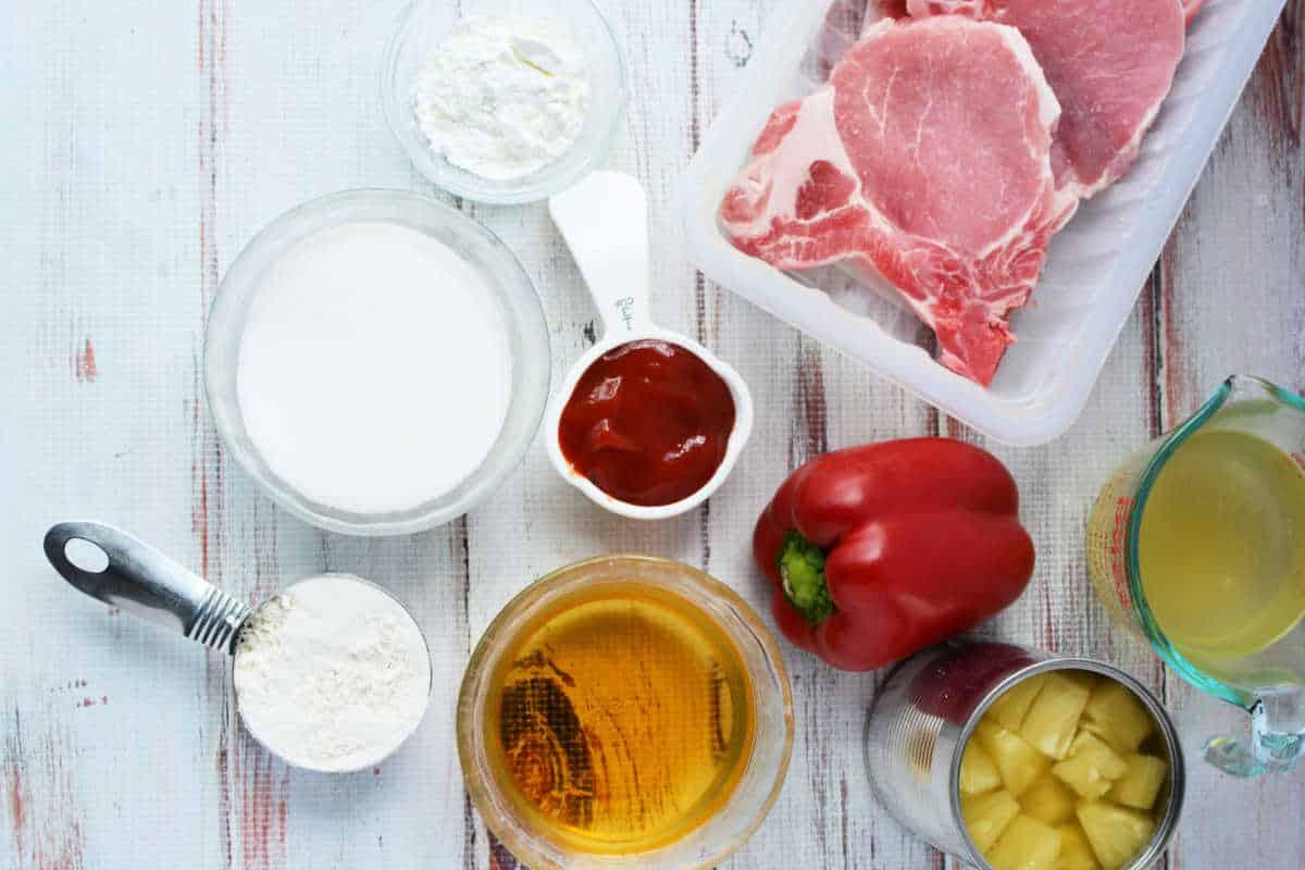 Raw pork chops, red bell pepper, flour, sugar, ketchup, pineapple, broth, and cornstarch sit on a white surface—ideal ingredients for delicious Pork Chop Recipes.