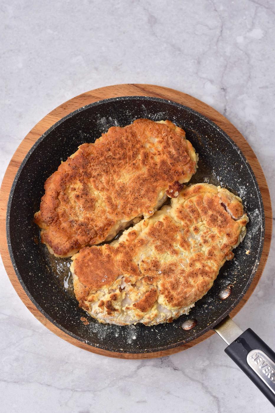 Two breaded pork cutlets and a succulent pork cube steak sizzle in a black pan set on a wooden board.
