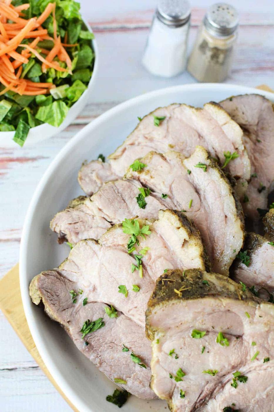 Sliced roast pork, tenderly prepared in an Instant Pot and garnished with fresh herbs, is elegantly arranged on a plate. In the background, a crisp salad accompanies the meal, alongside salt and pepper shakers.