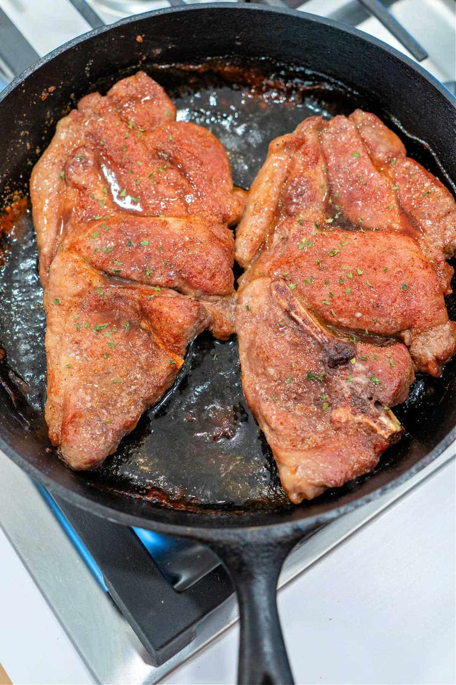 Two seasoned pork shoulder steaks sizzle in a black cast iron skillet on the stovetop.
