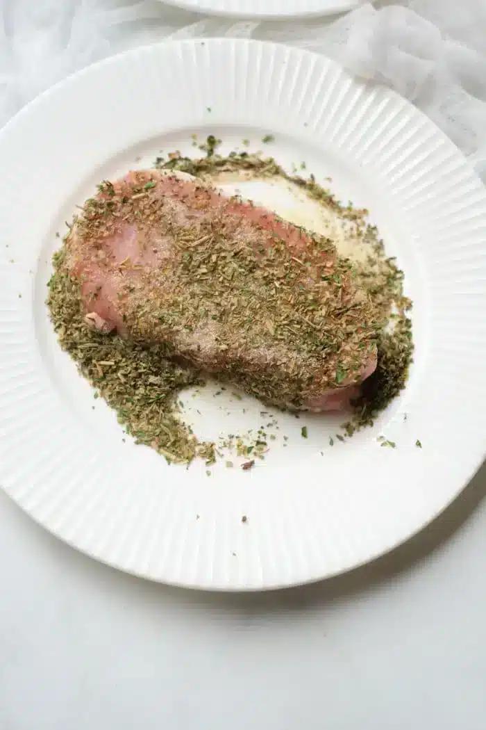 A pork steak, seasoned with a variety of herbs, rests on a white, ridged plate. The finely chopped herbs blanket the top of the meat and spill over onto the plate. Perfect for an air fryer, the background sets a light, textured scene.