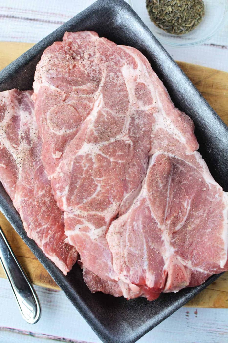 Raw pork steaks seasoned with pepper on a dark tray, ready for your next Pork Steak Recipe, with a bowl of dried herbs nearby.
