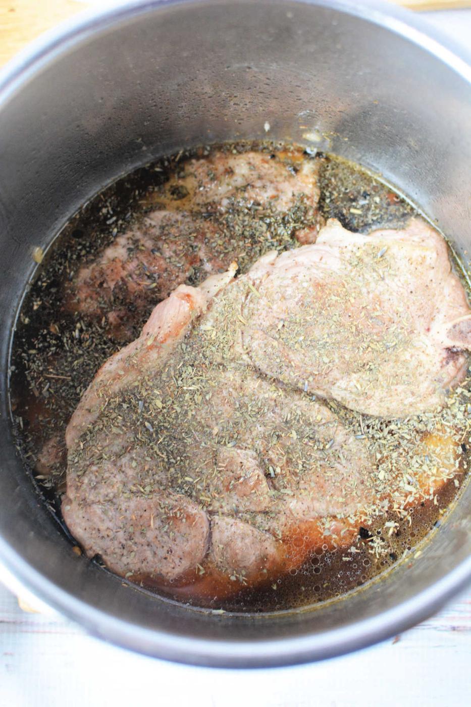 Two raw pork steaks with herbs and broth in a metal pot, ready for cooking—a simple start to a delicious pork steak recipe.