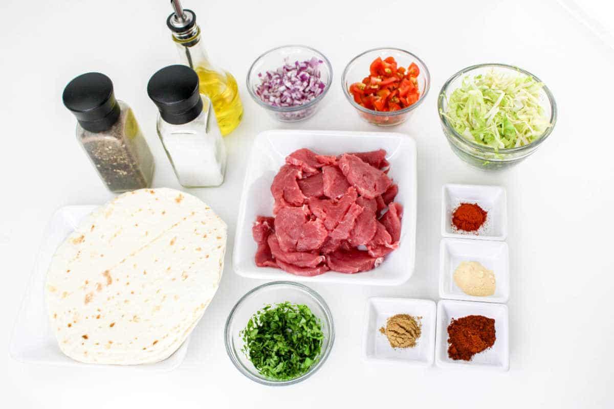 Ingredients for a meal are arranged on a white surface, including tortillas, raw pork slices, chopped cilantro, shredded cabbage, diced red onions, chopped red peppers, various spices in small dishes for making Pork Tenderloin Tacos, a bottle of olive oil, and salt and pepper shakers.