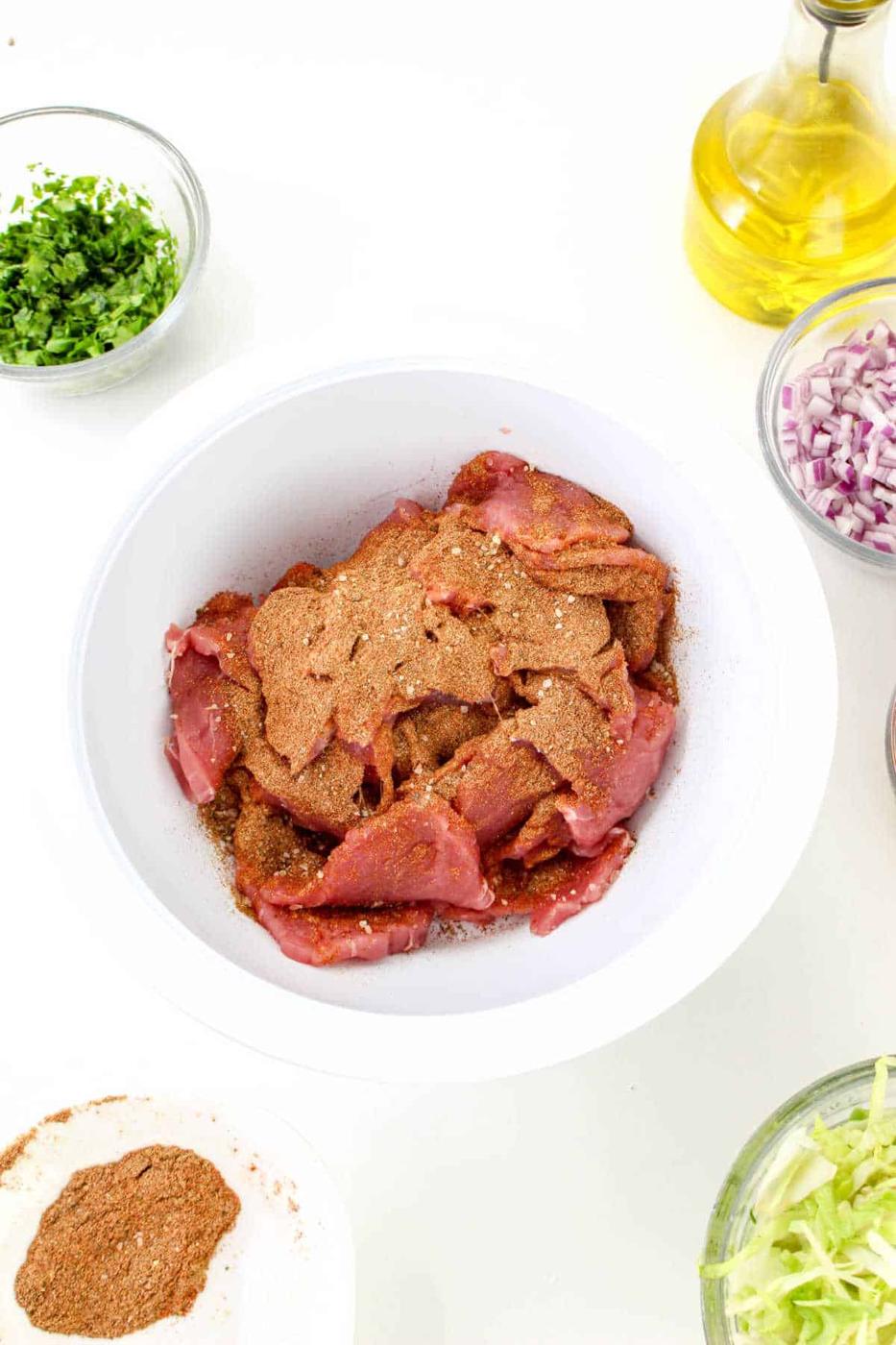 A white bowl contains raw pork pieces seasoned with a brown spice mix, surrounded by bowls of chopped herbs, diced red onions, shredded lettuce, a small bowl of additional spices resembling the ones used in pork tenderloin tacos, and a bottle of olive oil on a white background.