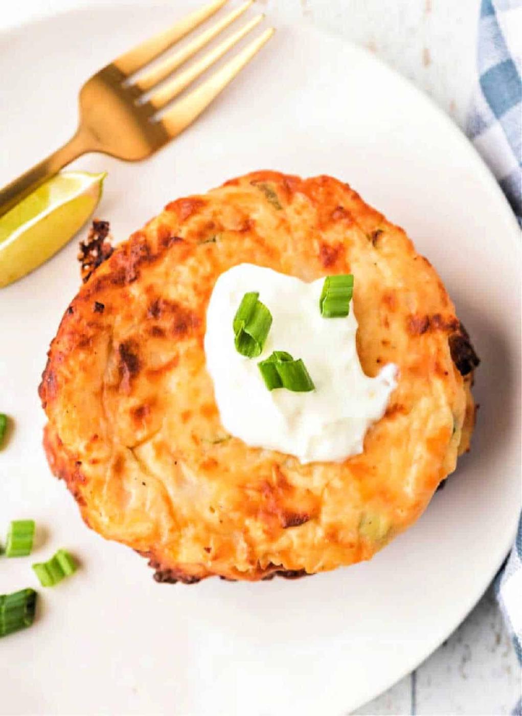 potato cake topped with sour cream and green onions on a white plate, alongside a lime wedge, fork, and an enticing dollop of fried mashed potatoes with cheese.