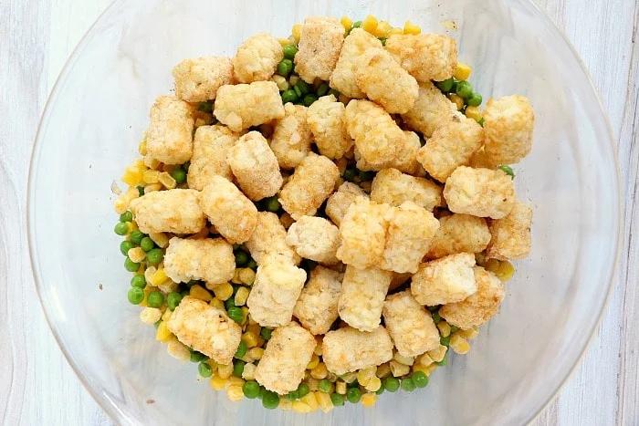A glass bowl filled with potato tot casserole with ground beef, peas, and corn, viewed from above.