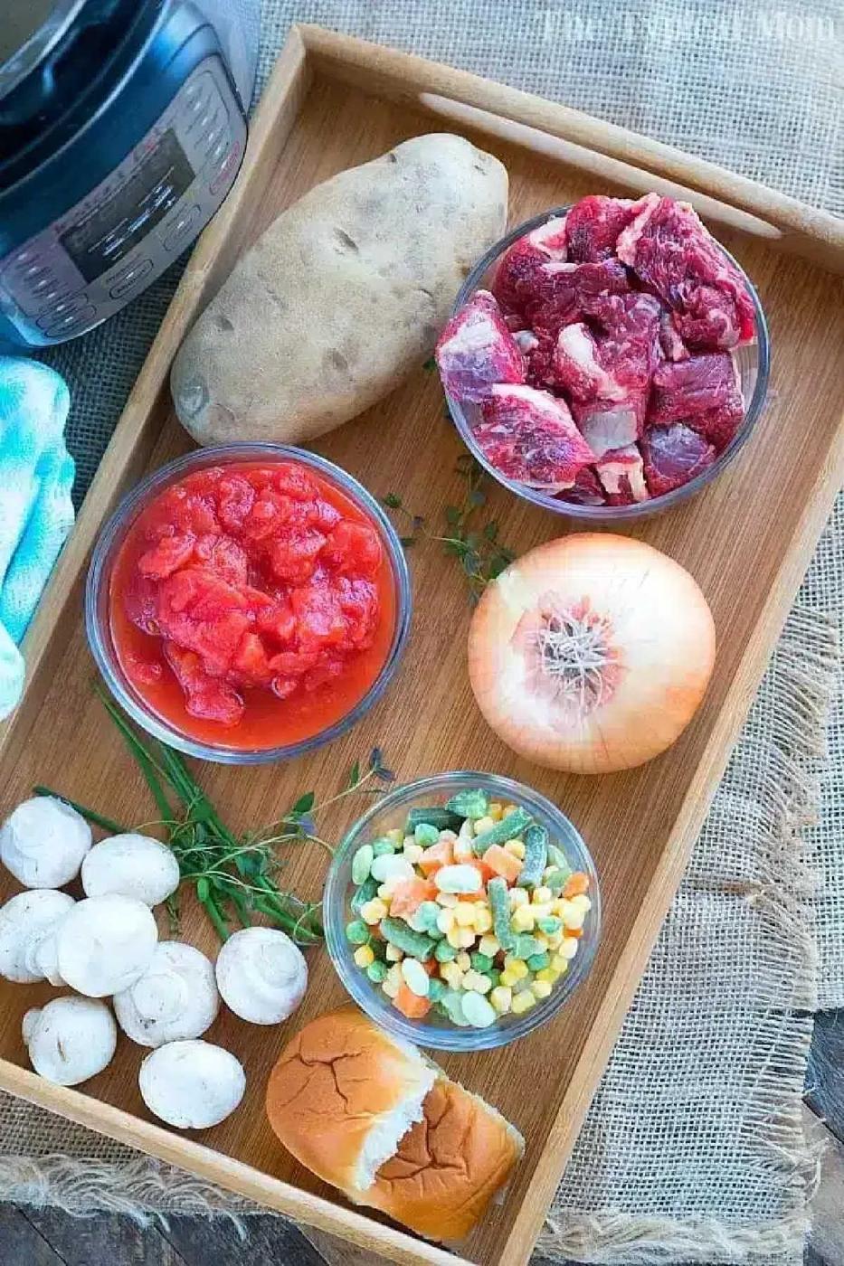 Tray with raw beef, diced tomatoes, potato, onion, mushrooms, mixed veggies, a bread roll, and an Instant Pot—everything you need to make a hearty pressure cooker beef pot pie soup.