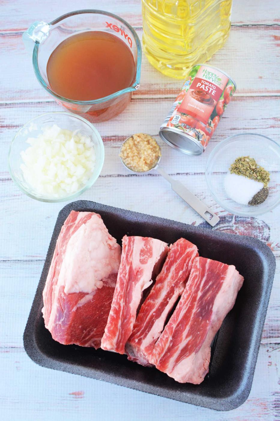 Ingredients for cooking: beef ribs, chopped onions, broth, tomato paste, minced garlic, spices, and oil on a table—perfect for a savory pressure cooker recipe.