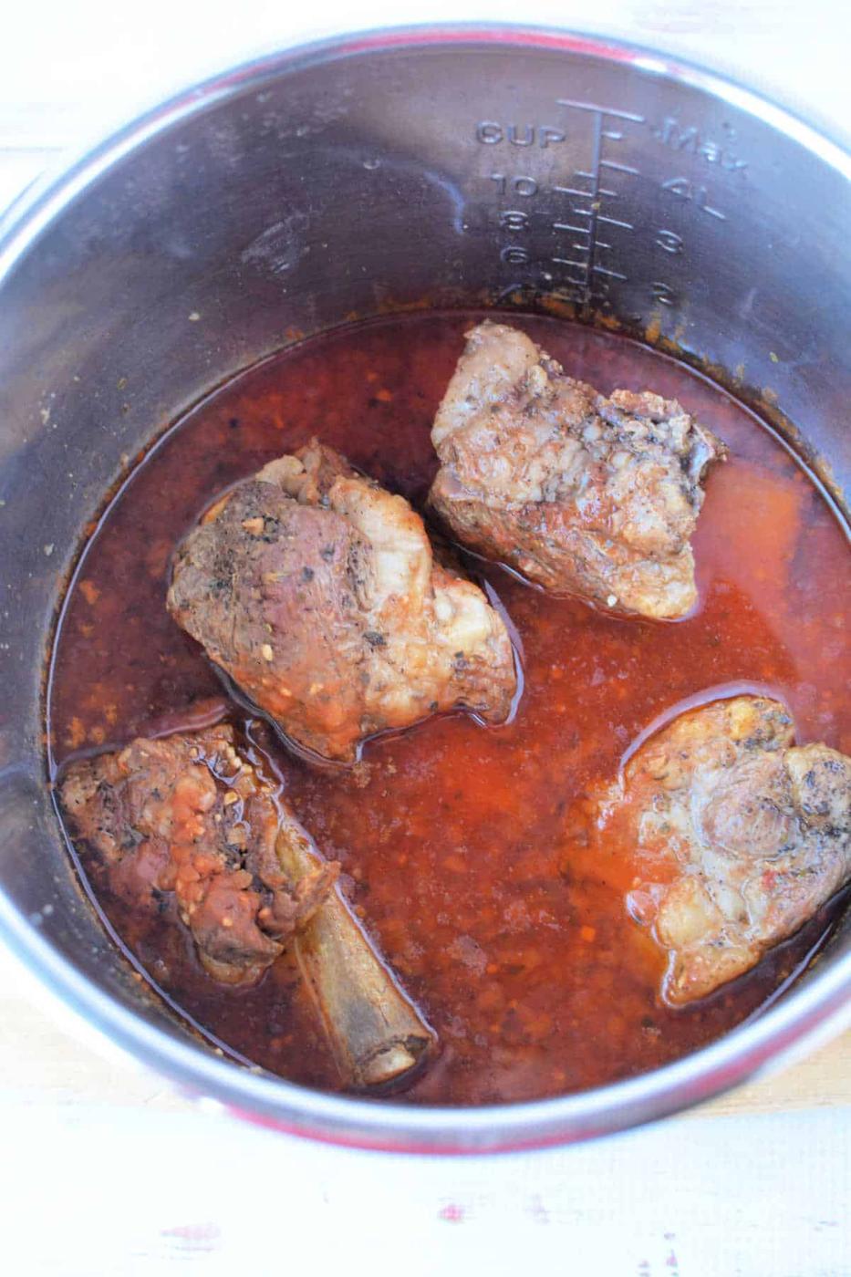 Cooked oxtail in a pot with rich, red broth, hinting at the tenderness achieved by using a pressure cooker. Four pieces are visible alongside aromatic spices and herbs.