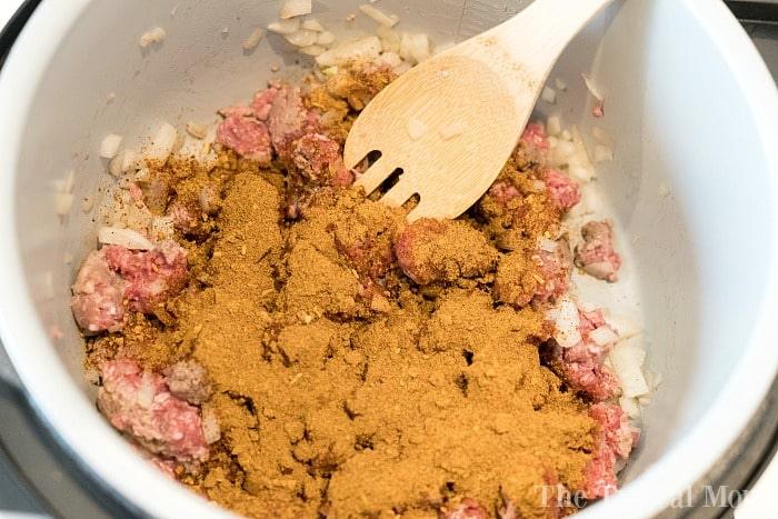 Chopped onions and ground meat are being sautéed in a pot with a wooden spoon. The ingredients are topped with a generous amount of ground spices, setting the stage for a delightful pressure cooker cheesy chili mac.
