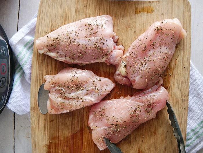 Four seasoned raw chicken thighs rest on a wooden cutting board with tongs nearby, ready to be transformed into a delectable instant pot meal