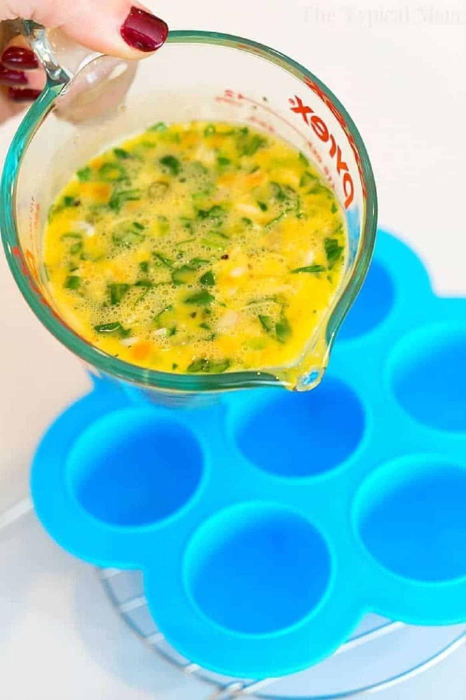 A hand pours egg mixture with vegetables from a measuring cup into a blue silicone mold to make pressure cooker egg bites.