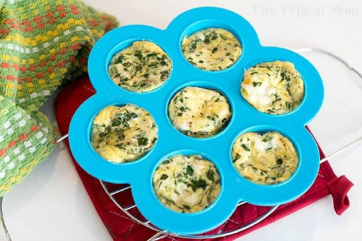 Seven pressure cooker egg bites with herbs in a blue silicone mold, resting on a red oven mitt and cooling rack.