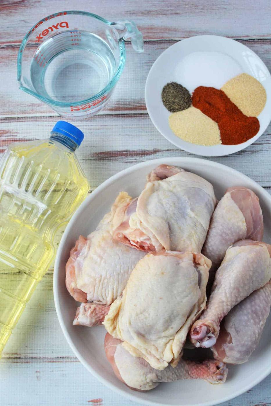 A raw chicken awaits transformation beside an oil bottle and a measuring cup of water, with a plate of spices ready to spice things up. Perfect ingredients for pressure cooker magic or crispy fried chicken bliss, all resting on a light wood surface.