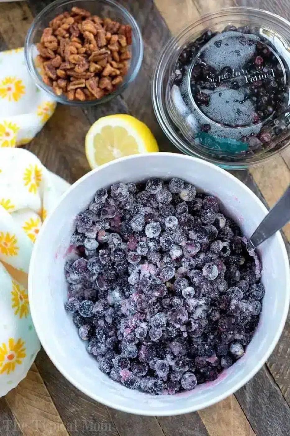 A bowl of frozen blueberries, a lemon half, a jar of pressure cooker jam, and a bowl of pecans rest on a wooden surface.