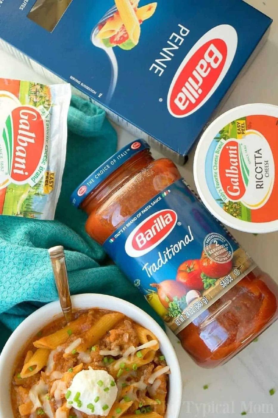 Penne pasta, ricotta cheese, and pasta sauce surround a bowl of pressure cooker lasagna casserole, topped with cheese and fresh herbs.