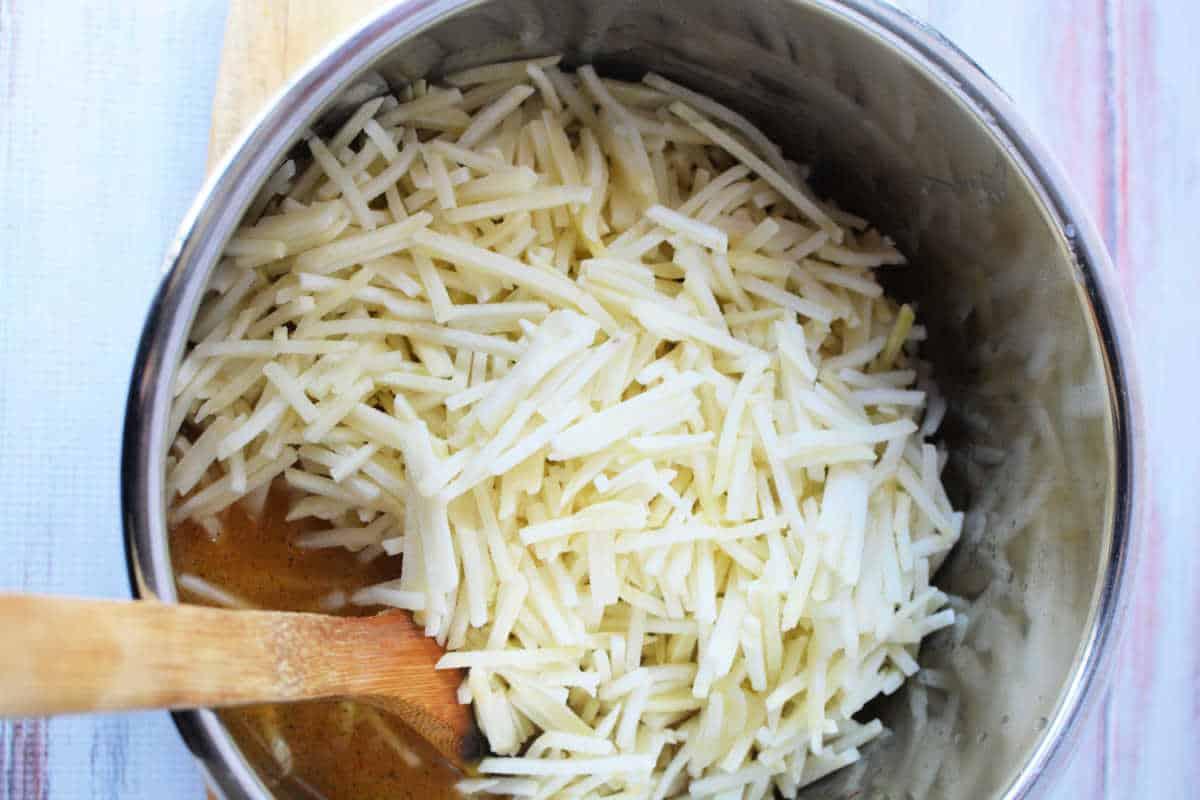 A pot filled with shredded potatoes, ready to become delicious frozen hash browns, and a wooden spoon rests on a light wooden surface.