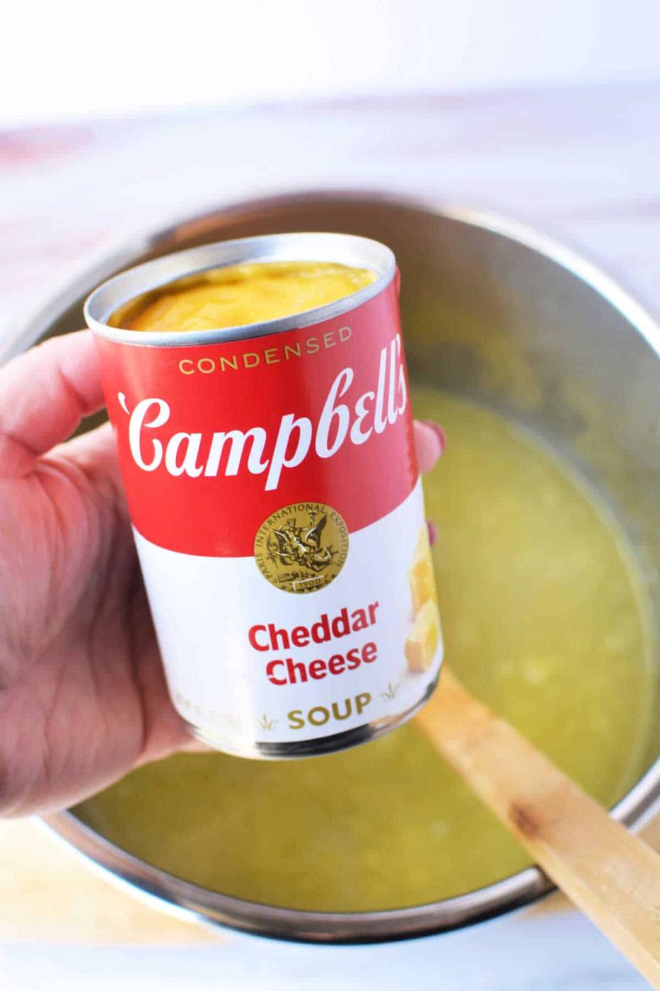 A hand holds a Campbells Cheddar Cheese soup can above a pot containing yellow potato soup with a wooden spoon, perfect for transforming with frozen hash browns in your pressure cooker.