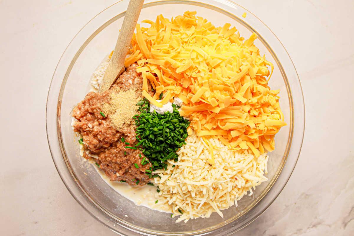 Ingredients in a bowl: shredded cheese, herbs, ground meat, and cream mix enticingly, ready to transform into savory sausage balls. A wooden spoon rests on the side, waiting to blend the flavors under the gentle guidance of a pressure cooker.