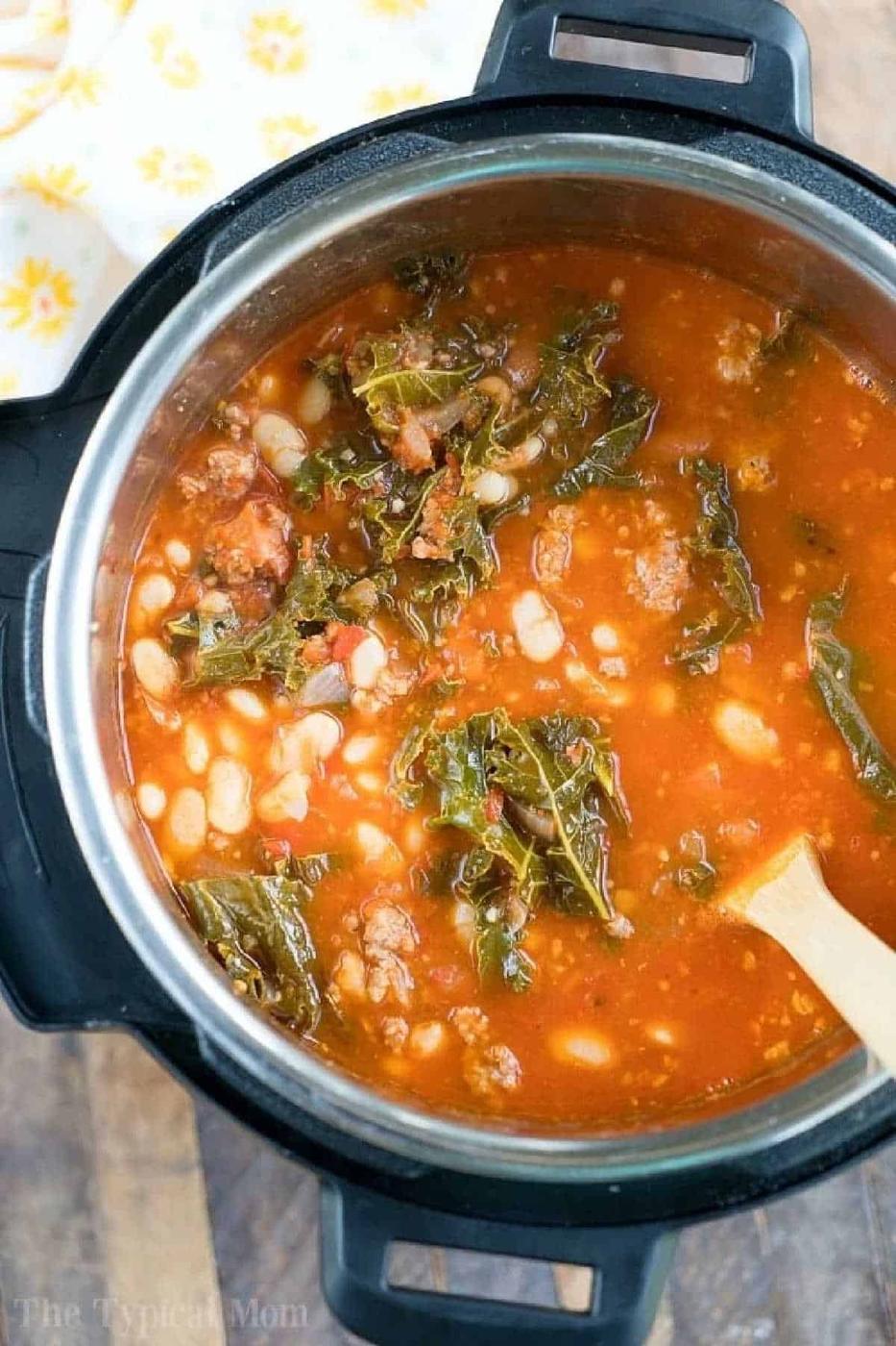 A pot of pressure cooker sausage soup with beans, leafy greens, and tomato broth is being stirred with a wooden spoon.