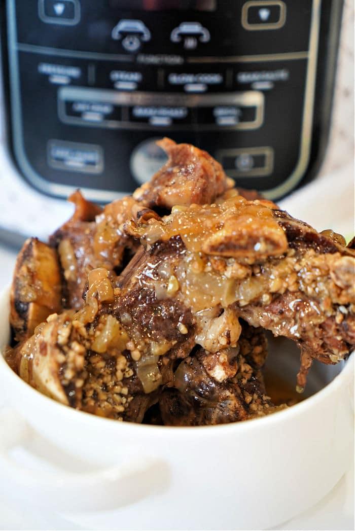 A bowl of tender short ribs with onions and spices sits elegantly in front of a pressure cooker, reminiscent of the savory allure found in short rib dishes.