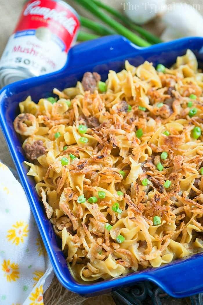 A blue baking dish brimming with noodle casserole topped with crispy onions and garnished with green peas. A can of Campbell's cream of mushroom soup and a bunch of green onions sit in the background, echoing a delicious twist reminiscent of pressure cooker stroganoff.