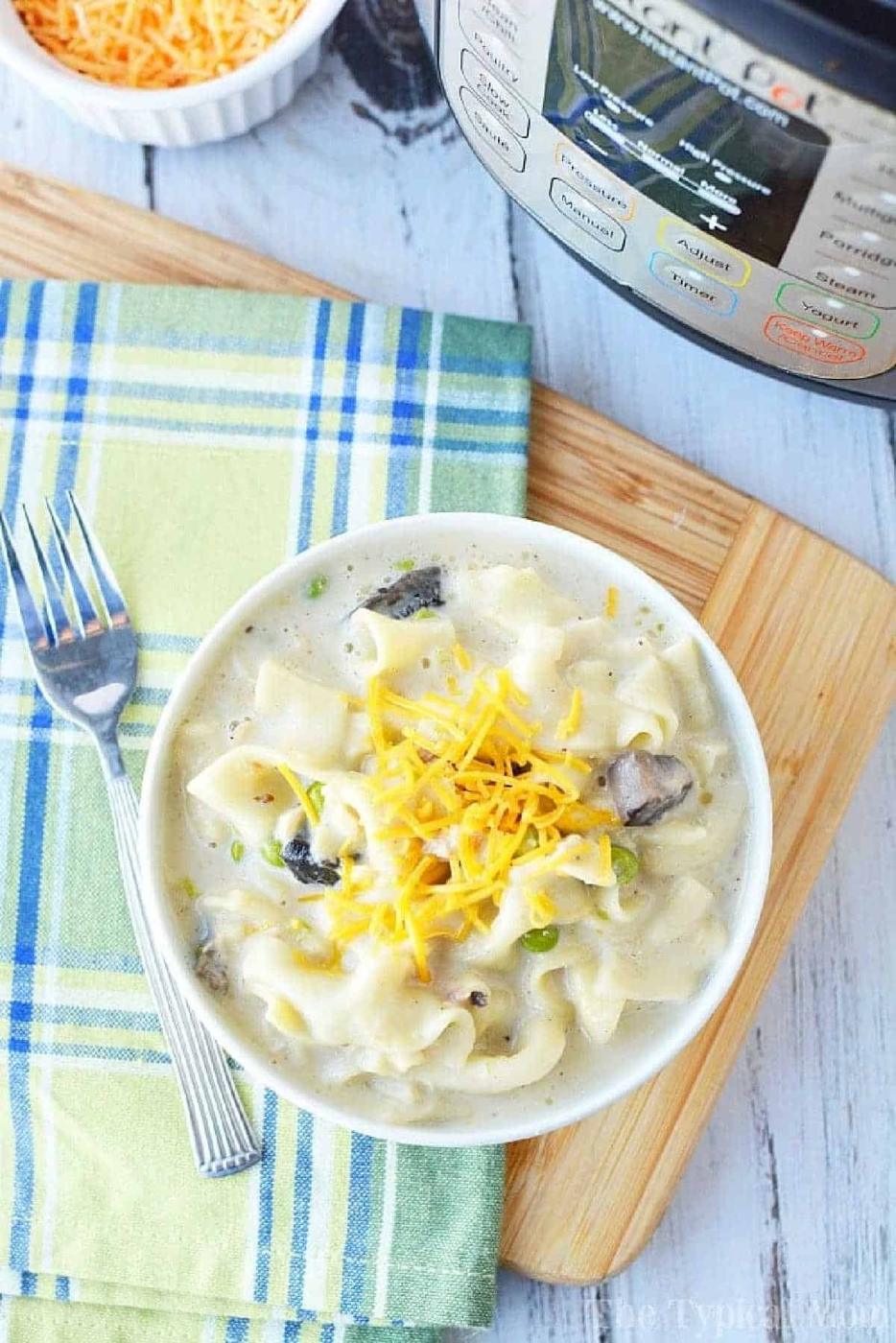 A bowl of creamy noodle soup inspired by pressure cooker tuna noodle casserole, topped with shredded cheese, sits next to a fork and an Instant Pot.