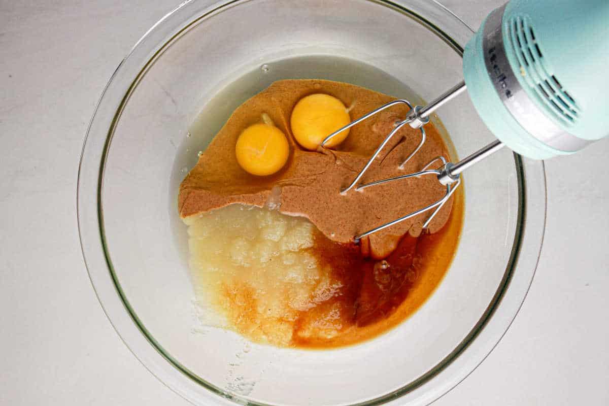 Mixer over a glass bowl with eggs, apple sauce, and batter ready to be mixed for a delicious protein brownie recipe.
