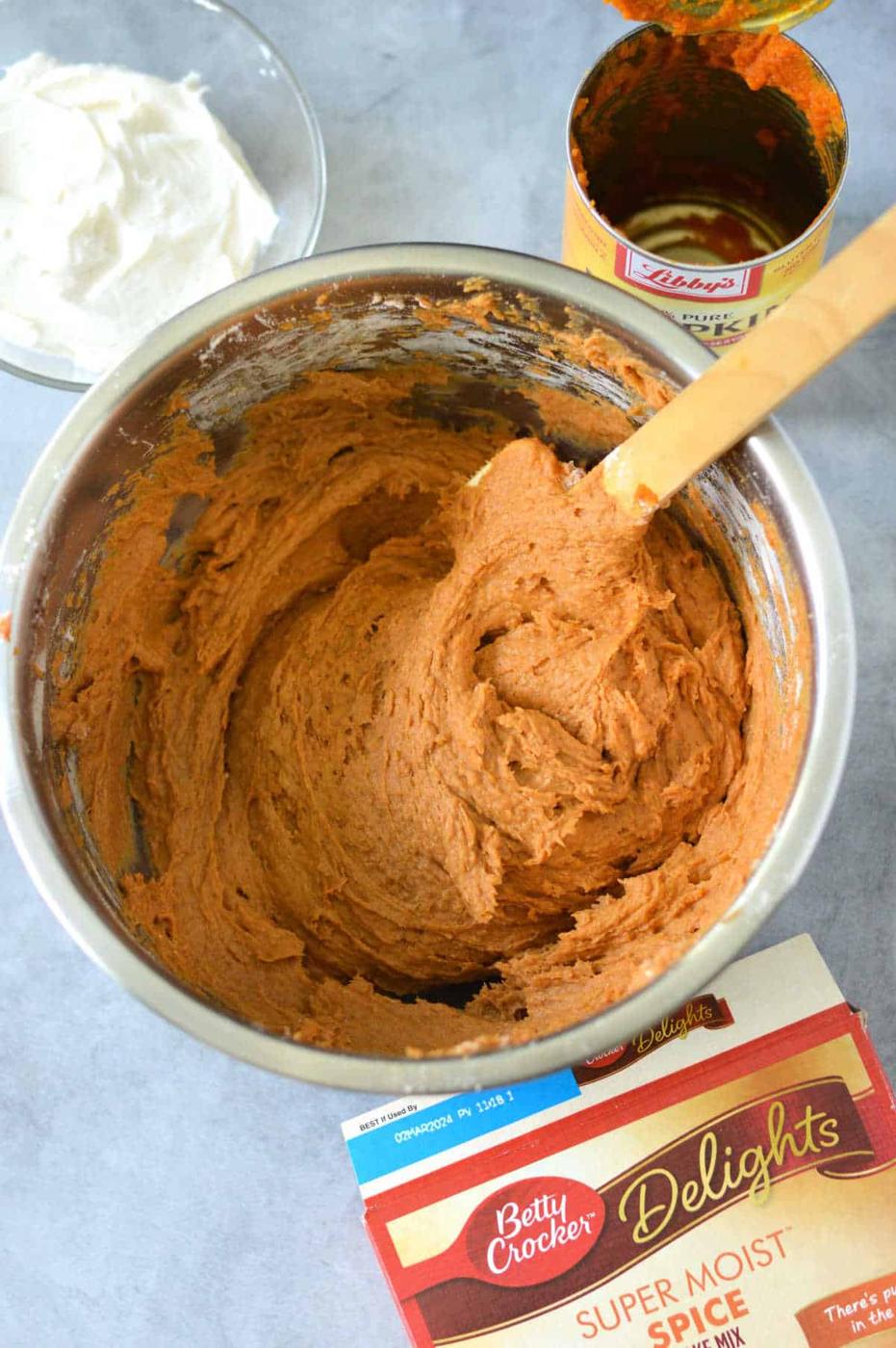 A mixing bowl filled with pumpkin cake bars batter sits on a gray surface, surrounded by canned pumpkin and a cake mix box, ready to whip up delicious pumpkin bars with cake mix. Nearby, whipped topping waits to add the finishing touch.