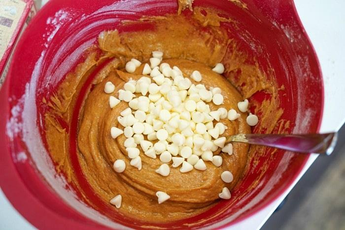 A red mixing bowl brimming with dough and white chocolate chips promises delightful pumpkin blondies, while a metal spoon rests inside, ready for action.