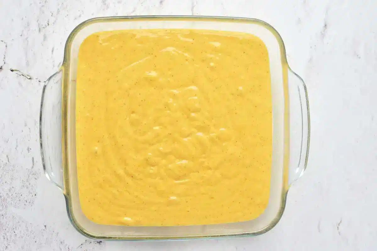 A square glass dish filled with a smooth, yellow batter sits on a light-colored marbled surface. The batter, perfect for pumpkin cake with yellow cake mix, has a few small bubbles scattered across the top, indicating that it has just been prepared and is ready to be baked.