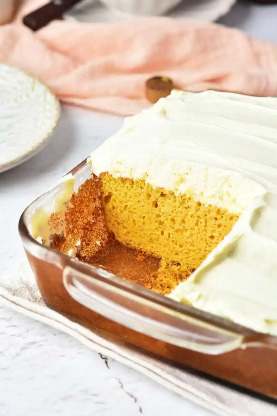 A rectangular glass baking dish contains a pumpkin cake with yellow cake mix, topped with white frosting. A square piece has been removed, revealing the cake's interior. The dish sits on a light-colored cloth, with a plate and peach-colored fabric in the background.