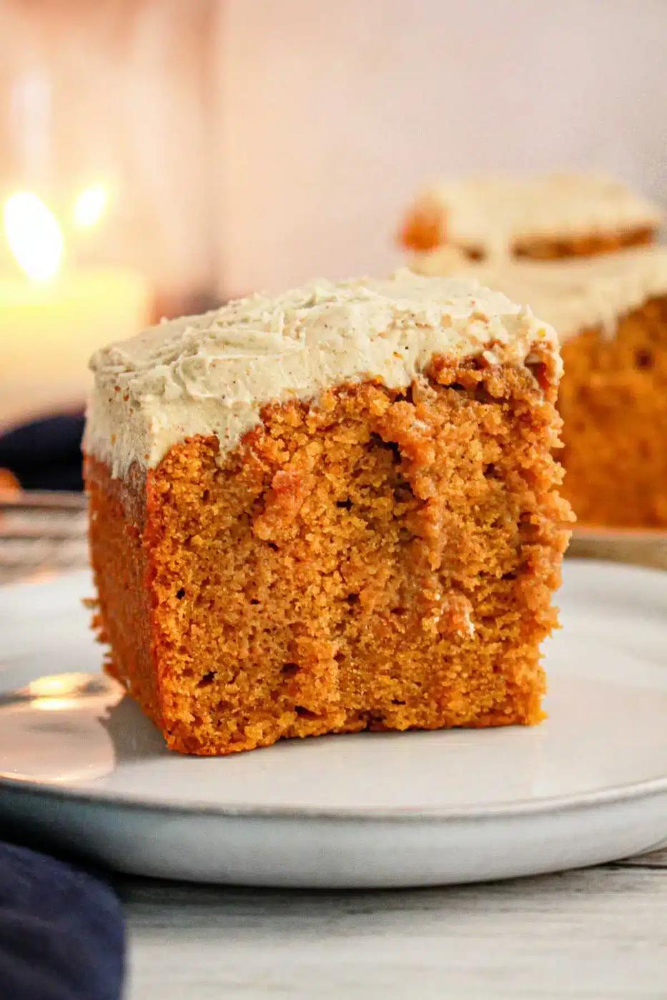 A close-up of a slice of Pumpkin Poke Cake with cream cheese frosting on a white plate. The cake has a golden-brown color and a moist texture, while the thick frosting covers the top of the slice. A blurred background shows another slice and a lit candle.