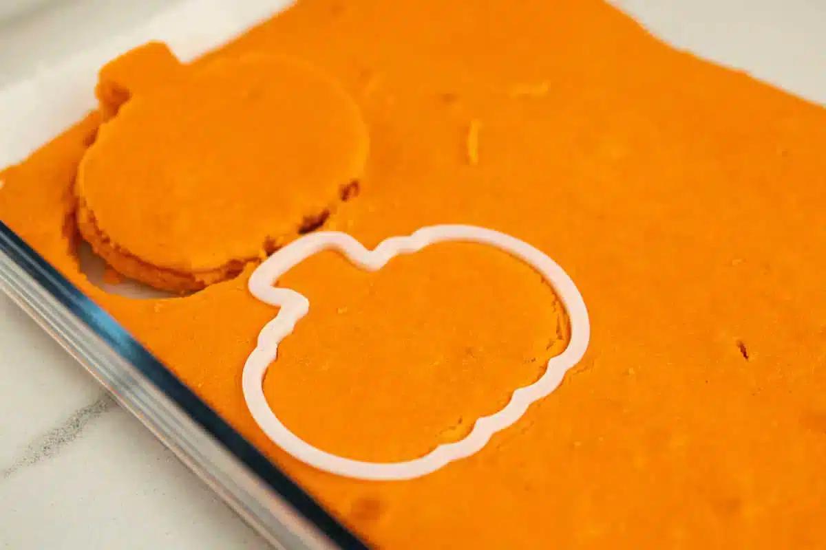 A sheet of orange dough with a pumpkin-shaped cookie cutter partially pressed into it. One pumpkin-shaped cut-out is already made and placed beside the cookie cutter. The scene, set on a white countertop, captures the playful process of creating festive shapes inside your kitchen.