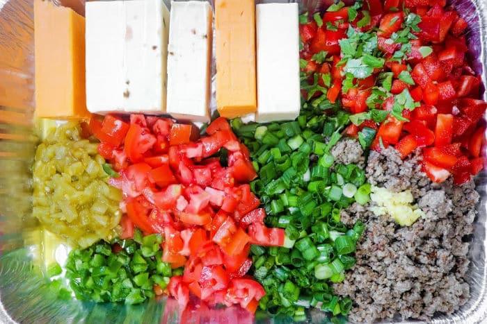 Chopped vegetables, ground meat, and cheese blocks arranged in sections in a foil tray—perfect for assembling a Queso Dip with Meat.