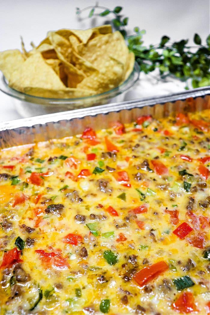 A cheesy ground beef casserole with chopped vegetables, inspired by Queso Dip with Meat, served next to a bowl of tortilla chips.