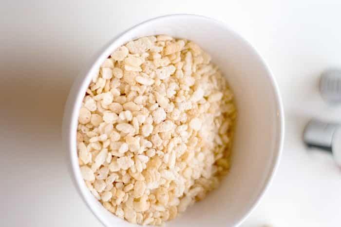 A white bowl filled with puffed rice cereal is placed on a white surface. The cereal is light, airy, and crisp, reminiscent of the texture in rice krispy treats. The edge of another object is slightly visible in the blurred background.