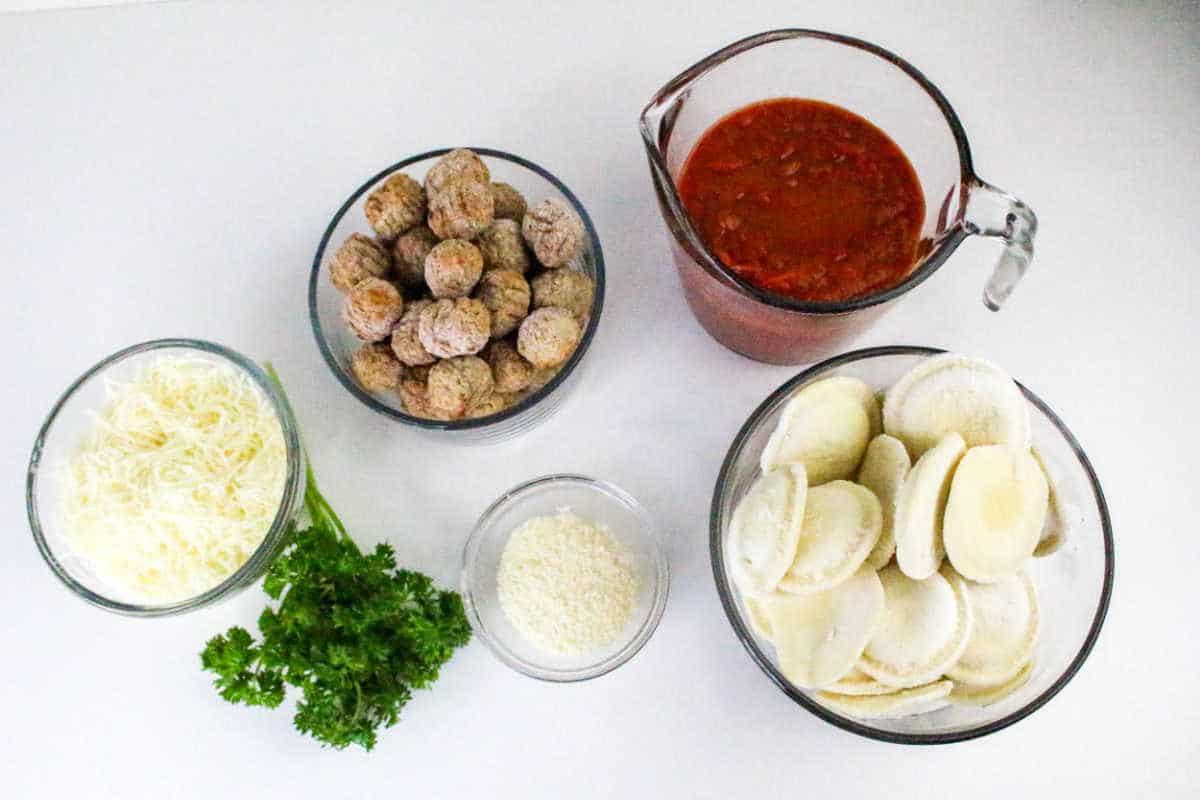 Bowls of shredded cheese, frozen meatballs, tomato sauce, ravioli, grated cheese, and fresh parsley on a white surface—everything you need to create a delicious Ravioli Casserole.