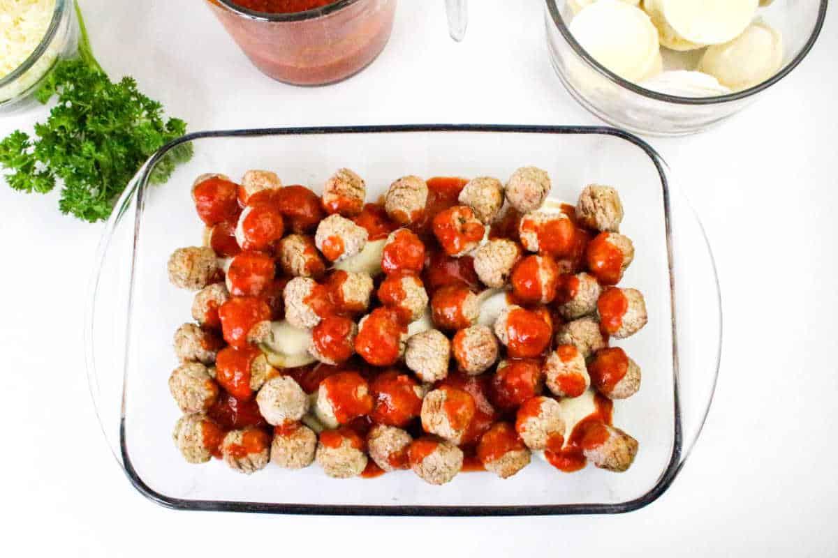 Glass baking dish with meatballs covered in tomato sauce, sliced mozzarella, and parsley on a white surface—perfect for serving alongside a homemade Ravioli Casserole.