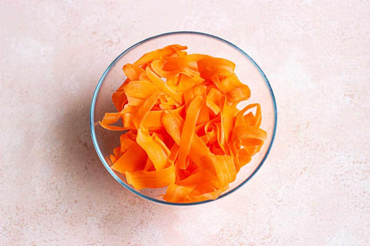 A glass bowl brimming with raw carrot salad featuring vibrant orange ribbons, set against a light beige background.