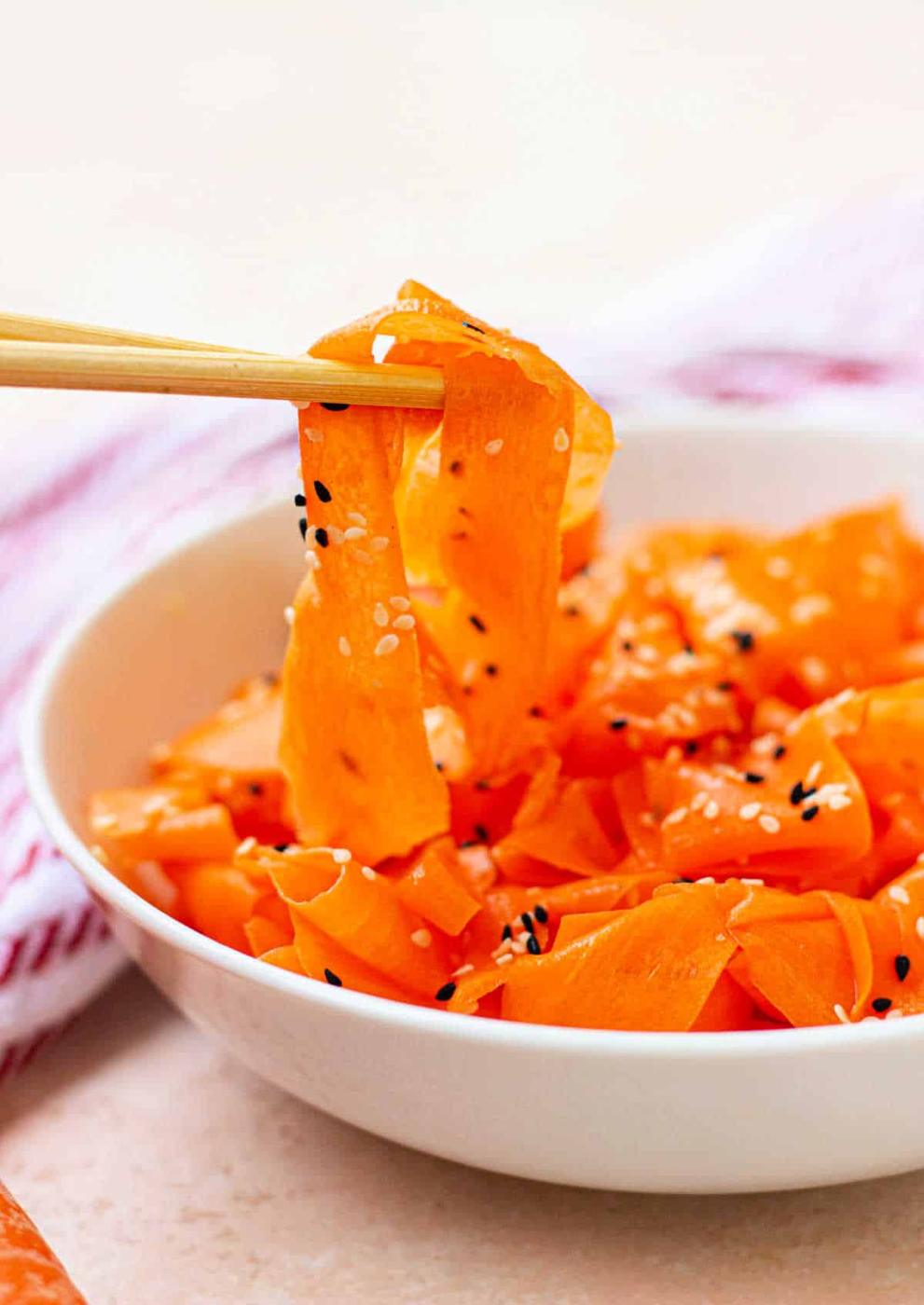 Chopsticks gracefully hold pieces of a delicious Raw Carrot Salad, sprinkled with sesame seeds, freshly lifted from a pristine white bowl.