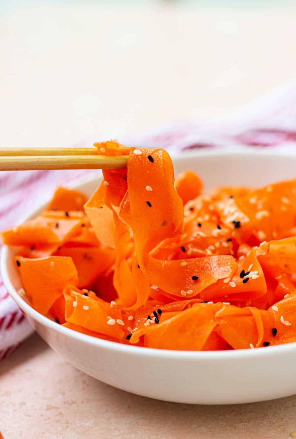 A bowl of raw carrot salad featuring delicate ribbons sprinkled with sesame seeds, gracefully lifted with chopsticks.