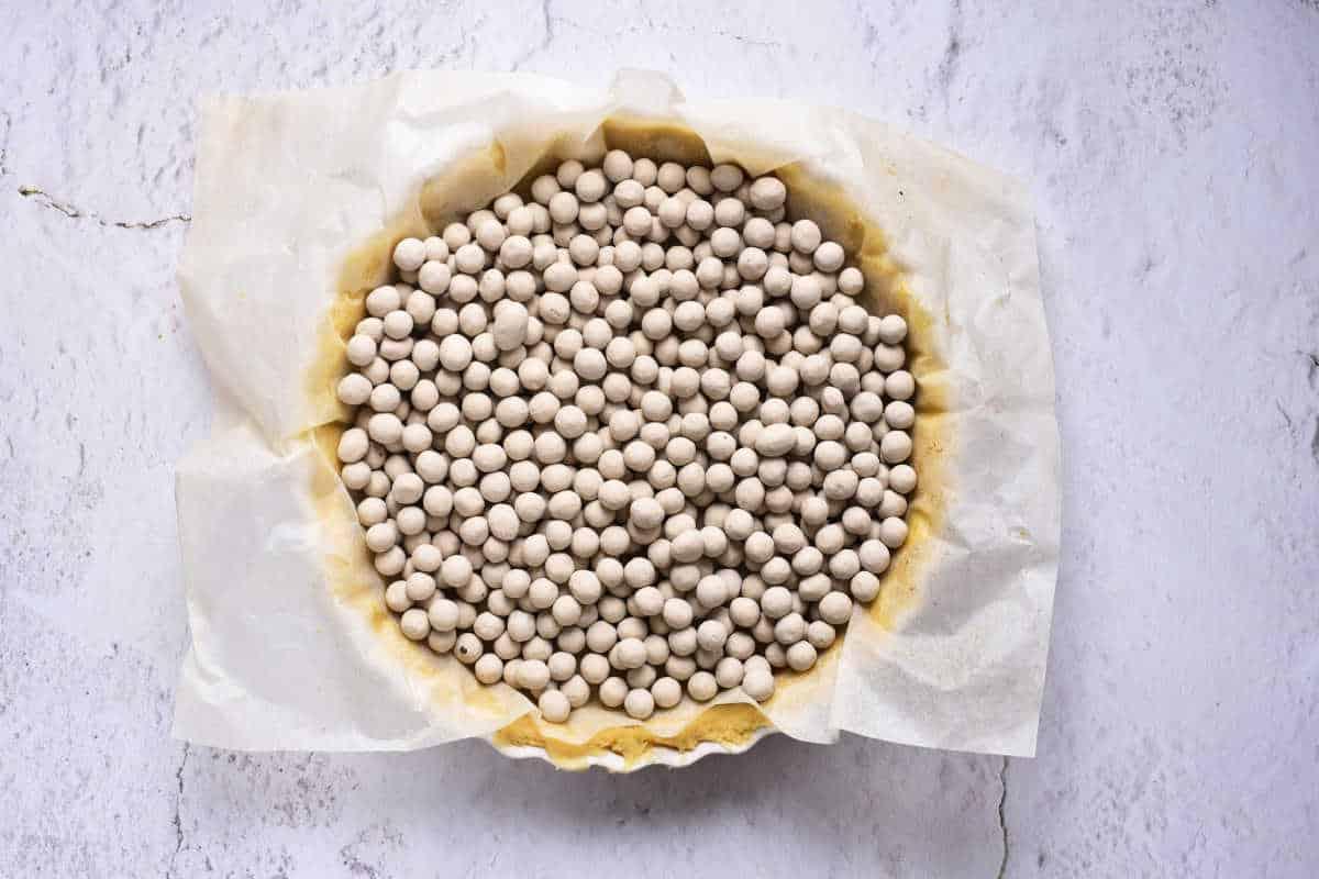 A round pie crust covered with parchment paper is filled with numerous small ceramic baking beads, preparing the foundation for a savory sweet potato pie with sweetened condensed milk. The pie is placed on a white, slightly textured surface.