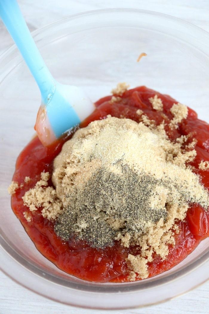 A clear mixing bowl contains ingredients for a savory sauce—ketchup, brown sugar, onion powder, and black pepper. A blue spatula rests in the bowl amid the partially combined mixture. Perfect for slathering on ribs destined for the Ninja air fryer.