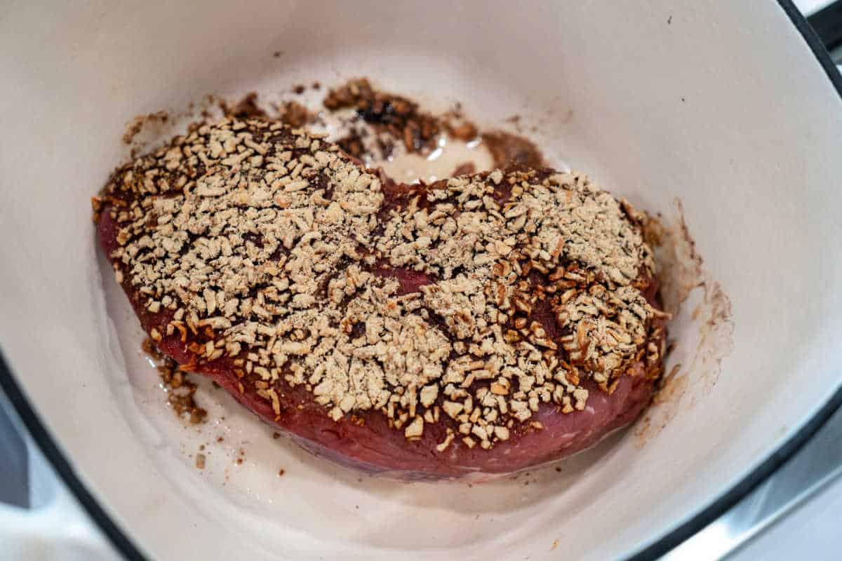 A roast piece of meat in a pot, topped with a mix of spices, herbs, and caramelized onions, ready to be cooked.