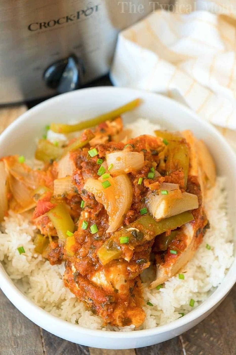 A bowl of white rice topped with a saucy chicken, onion, and bell pepper dish, made using an easy salsa chicken crockpot recipe, with a slow cooker in the background.