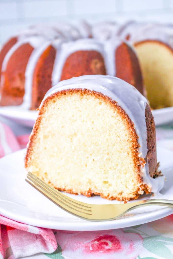 A slice of million dollar Pound Cake glazed bundt cake sits on a white plate with a gold fork, with the rest of the delicious cake in the background.