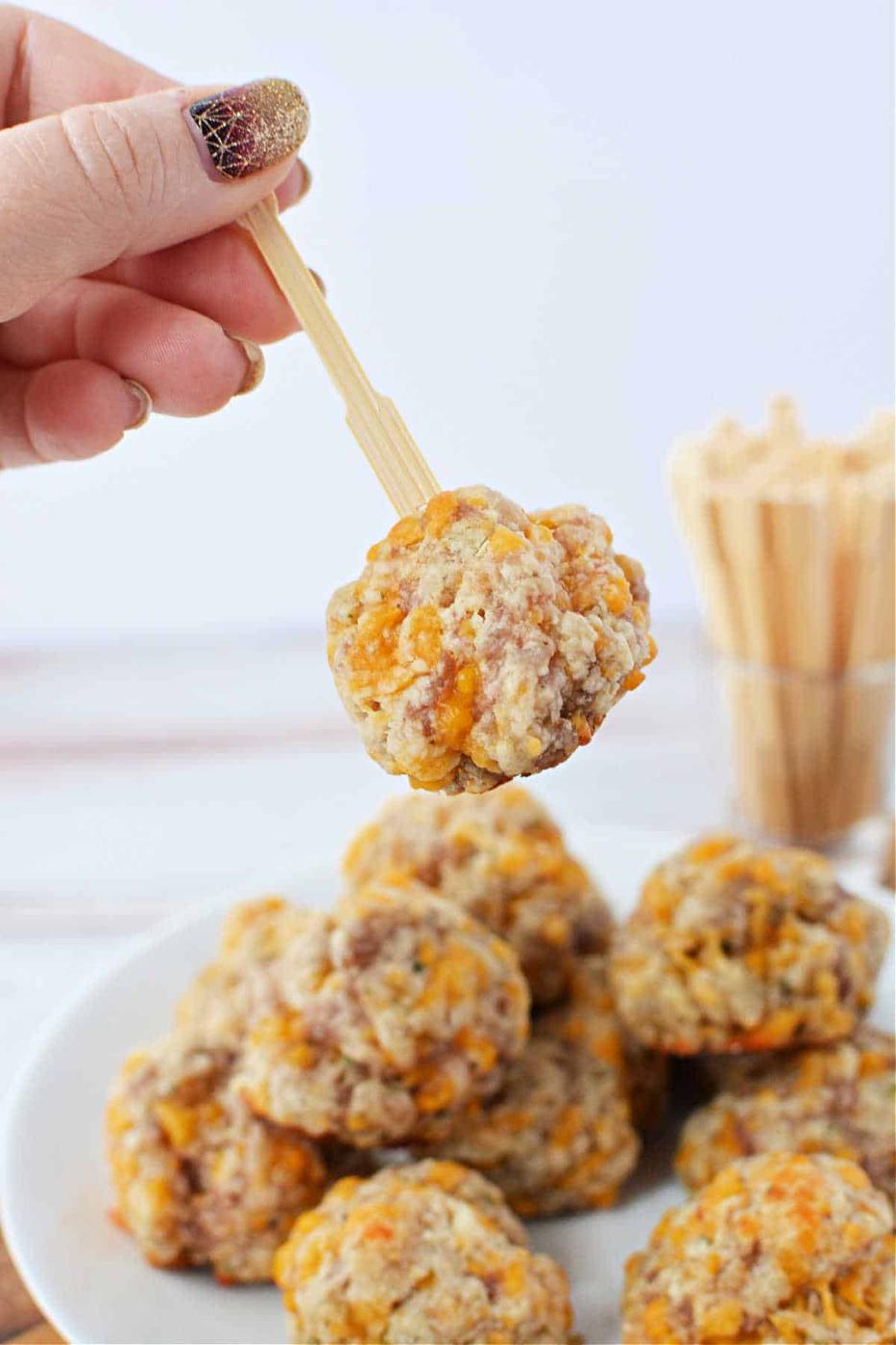 A hand holds a wooden pick with a cheese and sausage ball above a plate of delectable creations made using Red Lobster Biscuit Mix, enhancing the savory flavors of classic sausage balls.