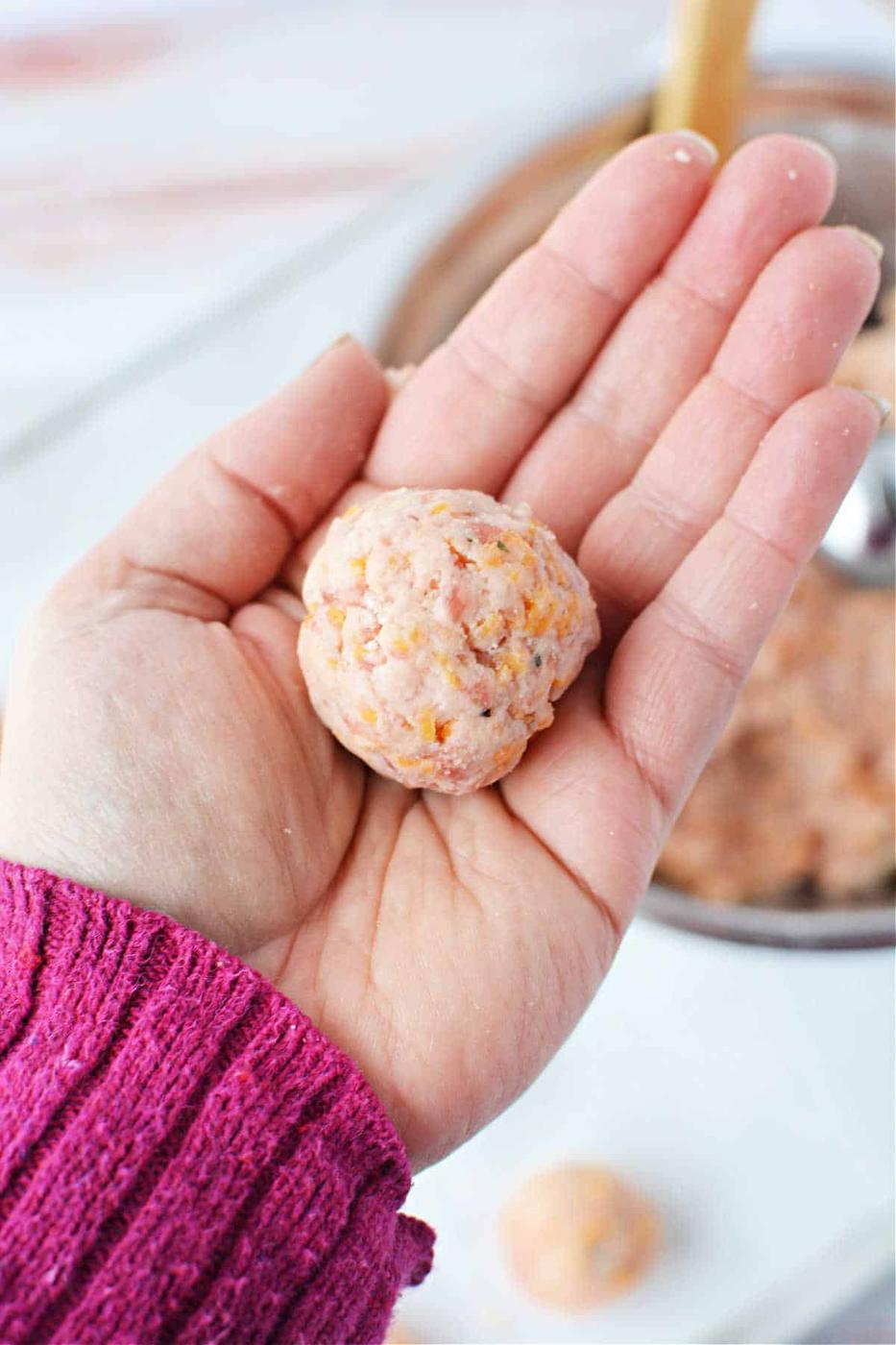 A hand wearing a pink sweater holds a round, raw sausage ball mixture above a table, with more of this savory blend nestled in the bowl below.